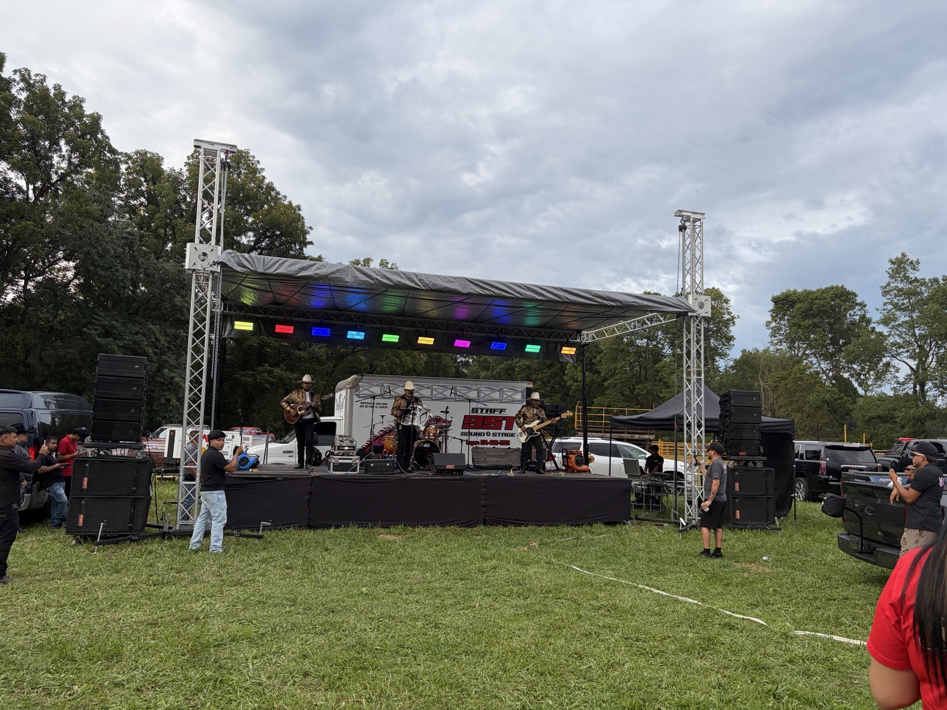 Músicos con sombreros tocan en un escenario con luces de colores y grandes altavoces frente a personas en el campo