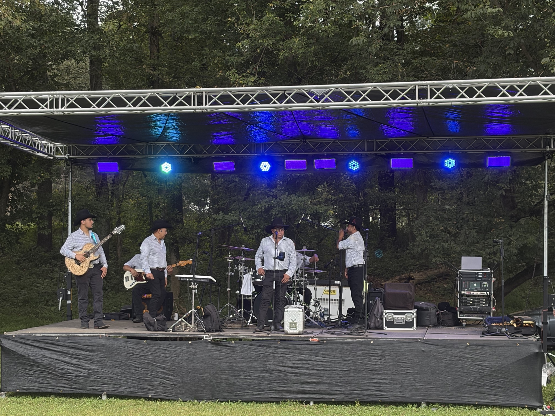 Músicos con sombreros tocan instrumentos en un escenario iluminado con luces azules y verdes rodeado de frondosos árboles